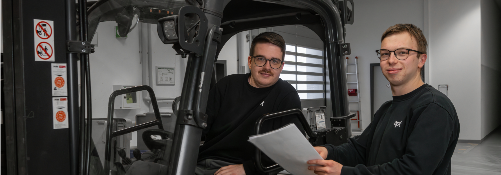 Auf dem Bild sind zwei Personen zu sehen, die in einem Gabelstapler sitzen und Unterlagen in der Hand halten.