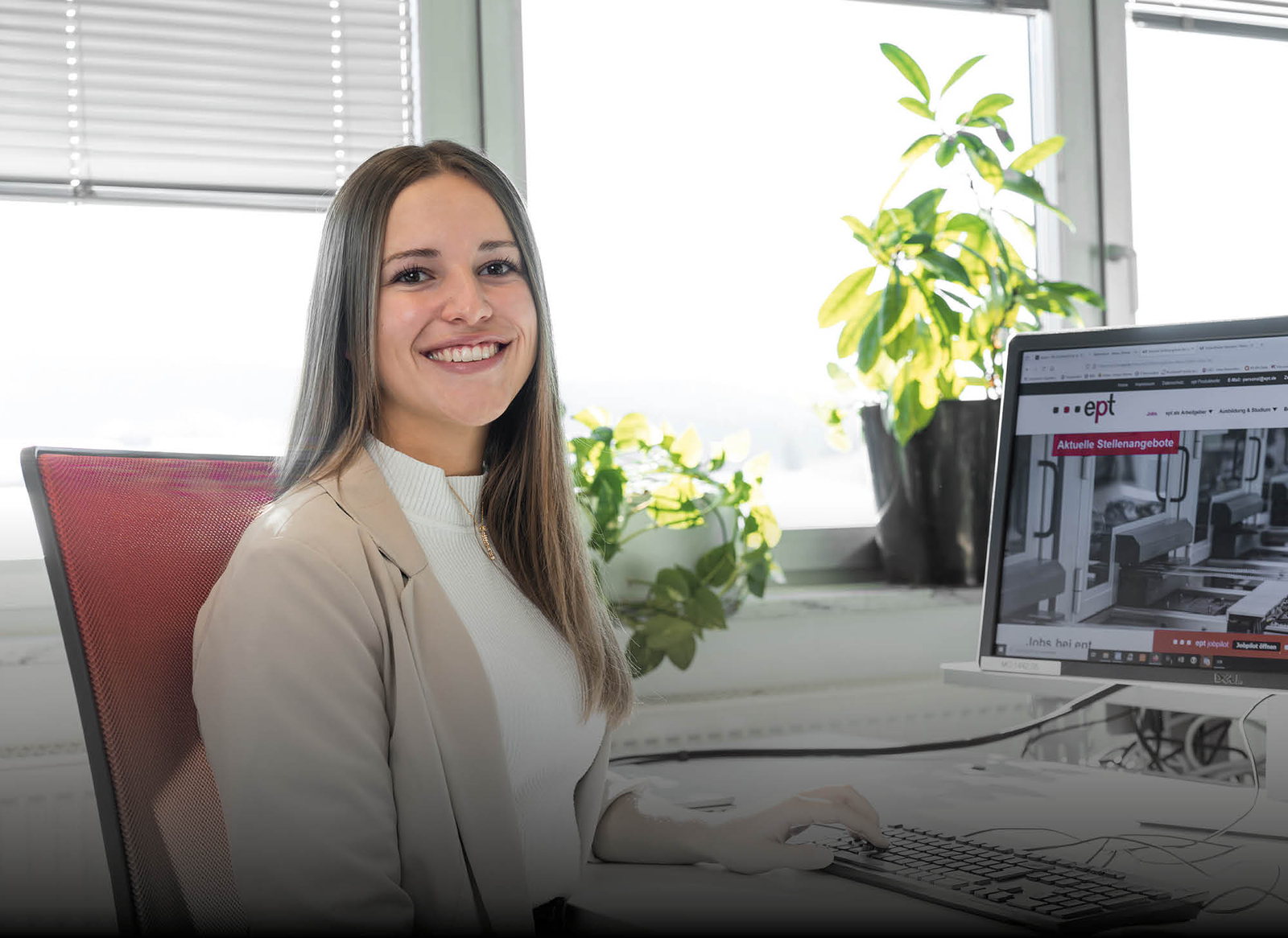 Auf dem Bild ist eine lächelnde Person zu sehen, die an einem Schreibtisch sitzt und vor einem Computer arbeitet, vor einem Fenster mit Pflanzen im Hintergrund.